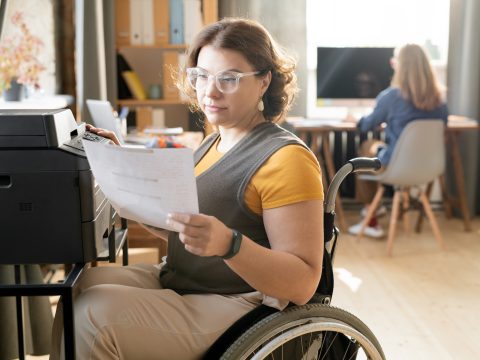 Young disable female office worker or secretary looking through paper while sitting by xerox machine and making copies against colleague