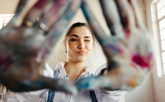 Carefree young painter framing her face with her hands. Happy female artist winking at the camera while standing in her art studio. Creative young woman making a frame with her colour painted hands.; Shutterstock ID 2104397717; purchase_order: purchase_order; job: job; client: client; other: other